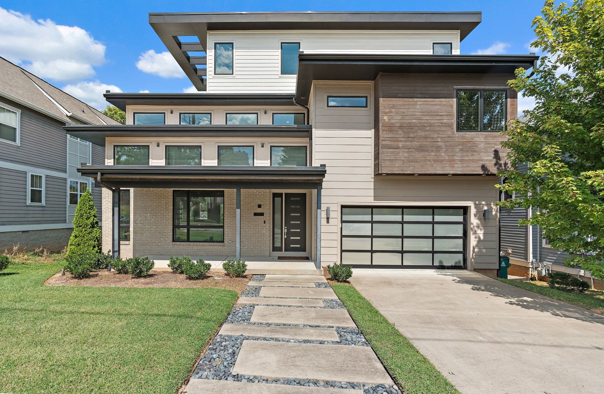 Front exterior of 1002 W Grove Avenue, a three story 2018 contemporary home with tan brick, light siding, stained wood panels, deep flat overhangs, a covered front porch, a glass panel garage door and a stone paver front walk, one of the 12 South homes for sale in Nashville
