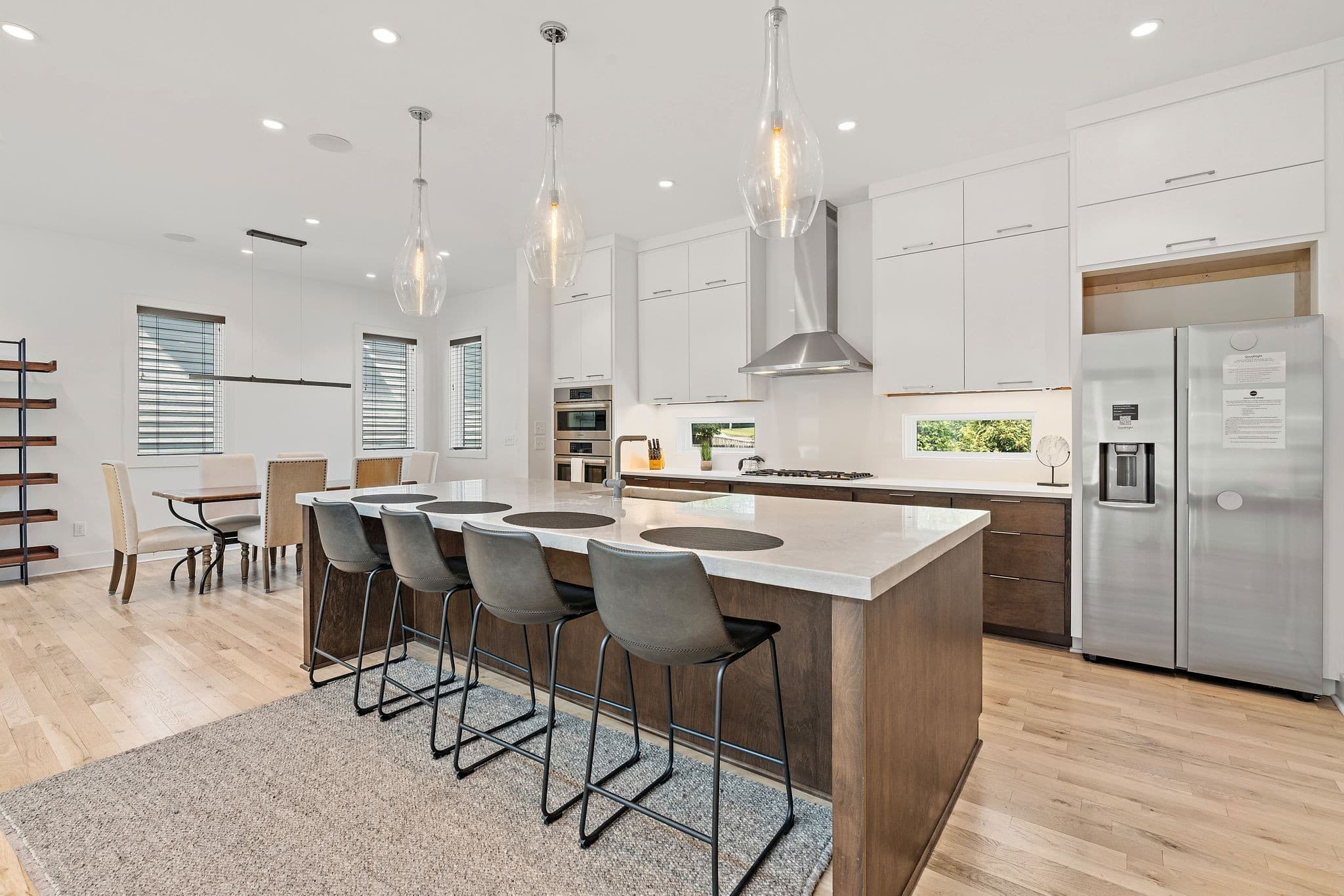 Open kitchen with a long island with four bar stools, wood lower cabinets, white upper cabinets to the ceiling, a stainless hood, glass pendant lights and a stainless refrigerator at 1002 W Grove Avenue in 12 South Nashville