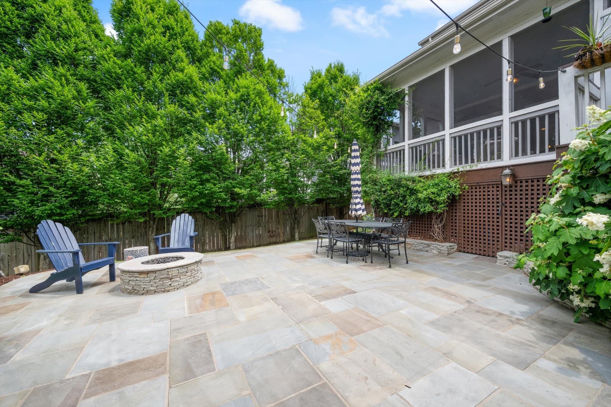 Fenced backyard with a stone patio, a round stone fire pit with two chairs, an outdoor dining table, string lights, and the screened porch above at 1008 Caldwell Avenue in 12 South Nashville