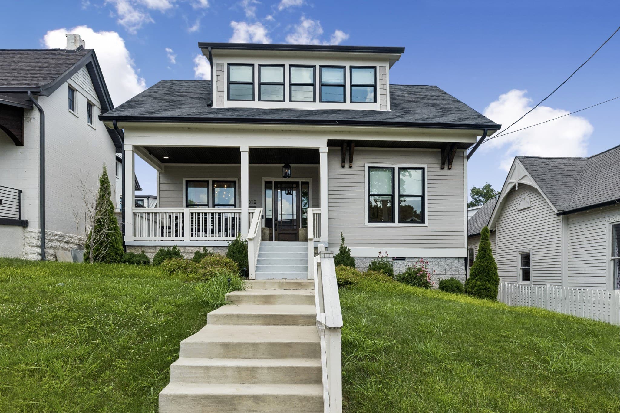 Street view of the front exterior of 1012 Lawrence Avenue, a light gray two story home with a covered front porch, white railings, a wide dormer with five windows and concrete steps rising up a sloped front lawn, one of the 12 South homes for sale in Nashville