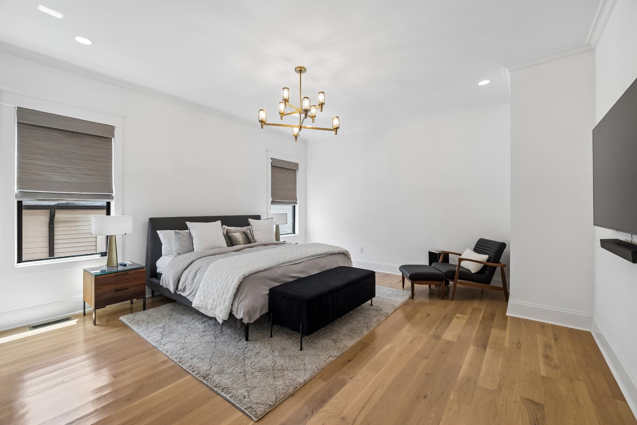Main level primary bedroom with light hardwood floors, a brass chandelier, woven window shades, an upholstered bed with a bench at its foot and a leather lounge chair at 1012 Lawrence Avenue in 12 South Nashville