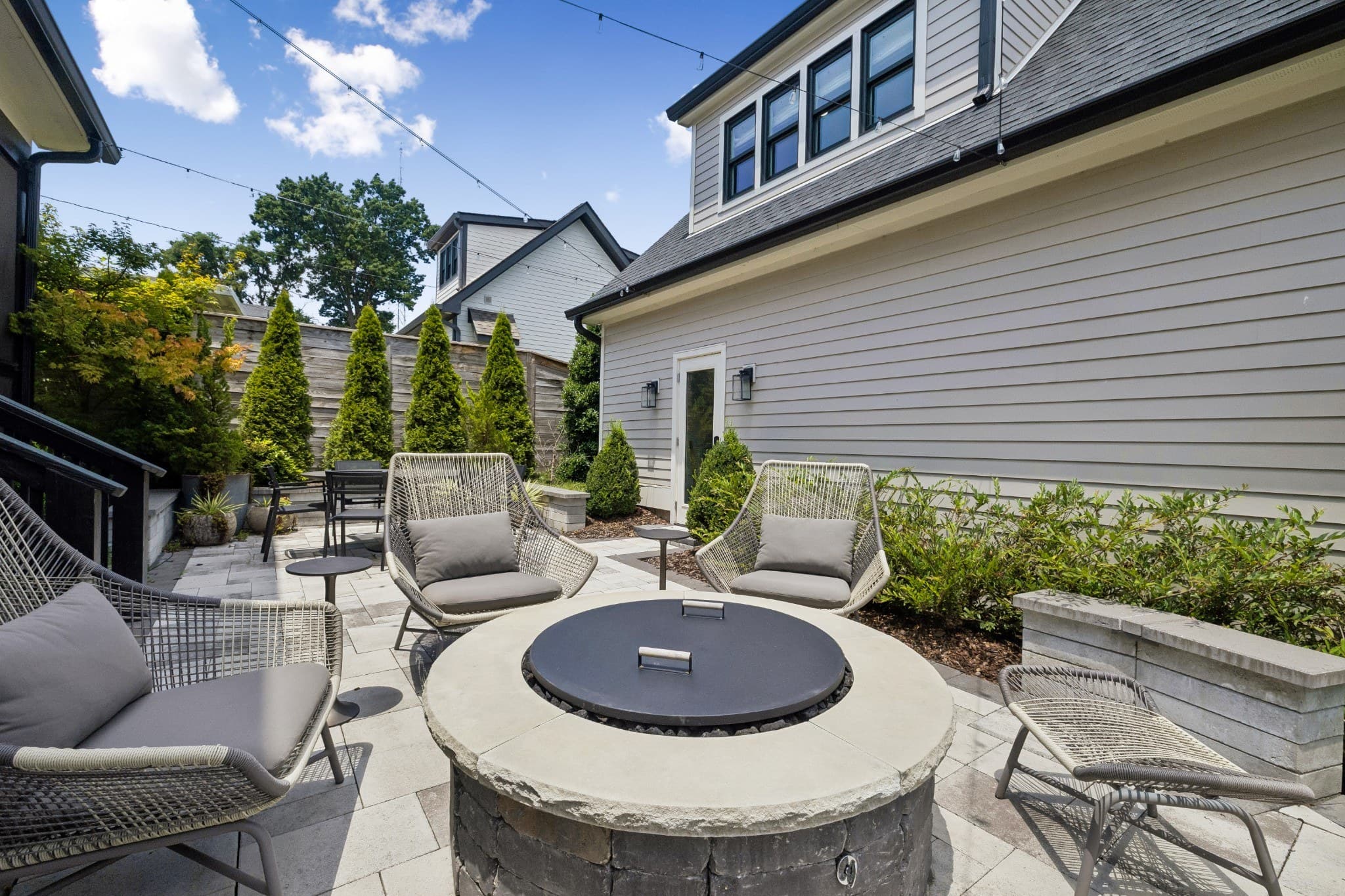 Private courtyard with a paver patio, a round stone gas fire pit with woven lounge chairs, string lights overhead and evergreens along a wood privacy fence at 1012 Lawrence Avenue in 12 South Nashville