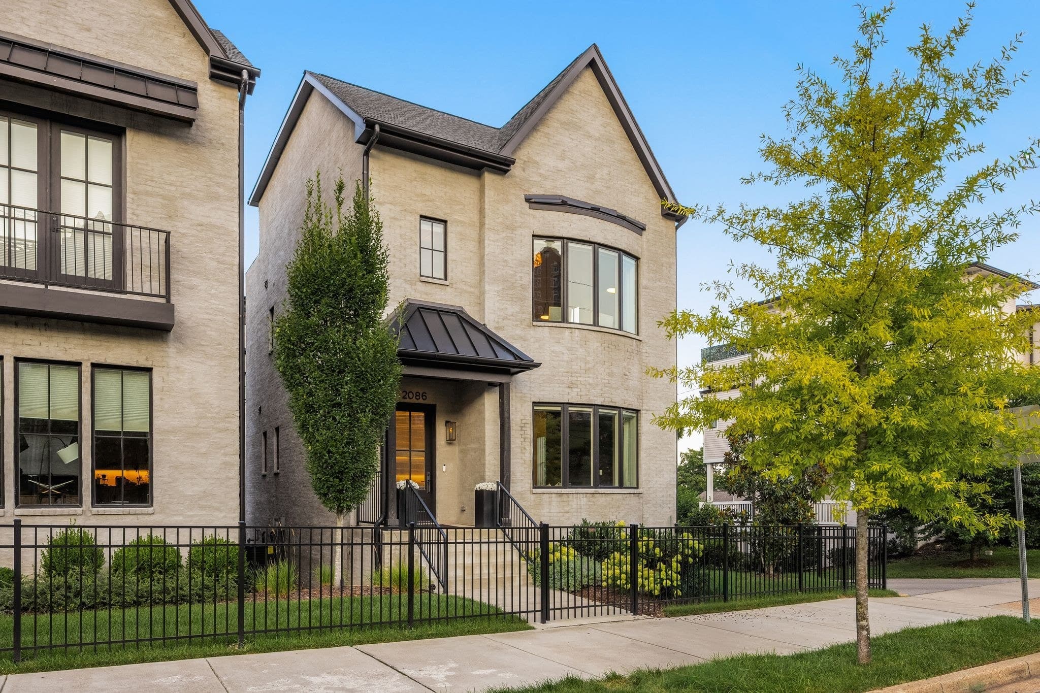 Street view of the front exterior of 2086 12th Avenue South, a cream brick greystone with the house number 2086 above the door, a black metal fence, a bay window, and a short walkway from the sidewalk, one of the 12 South homes for sale in Nashville