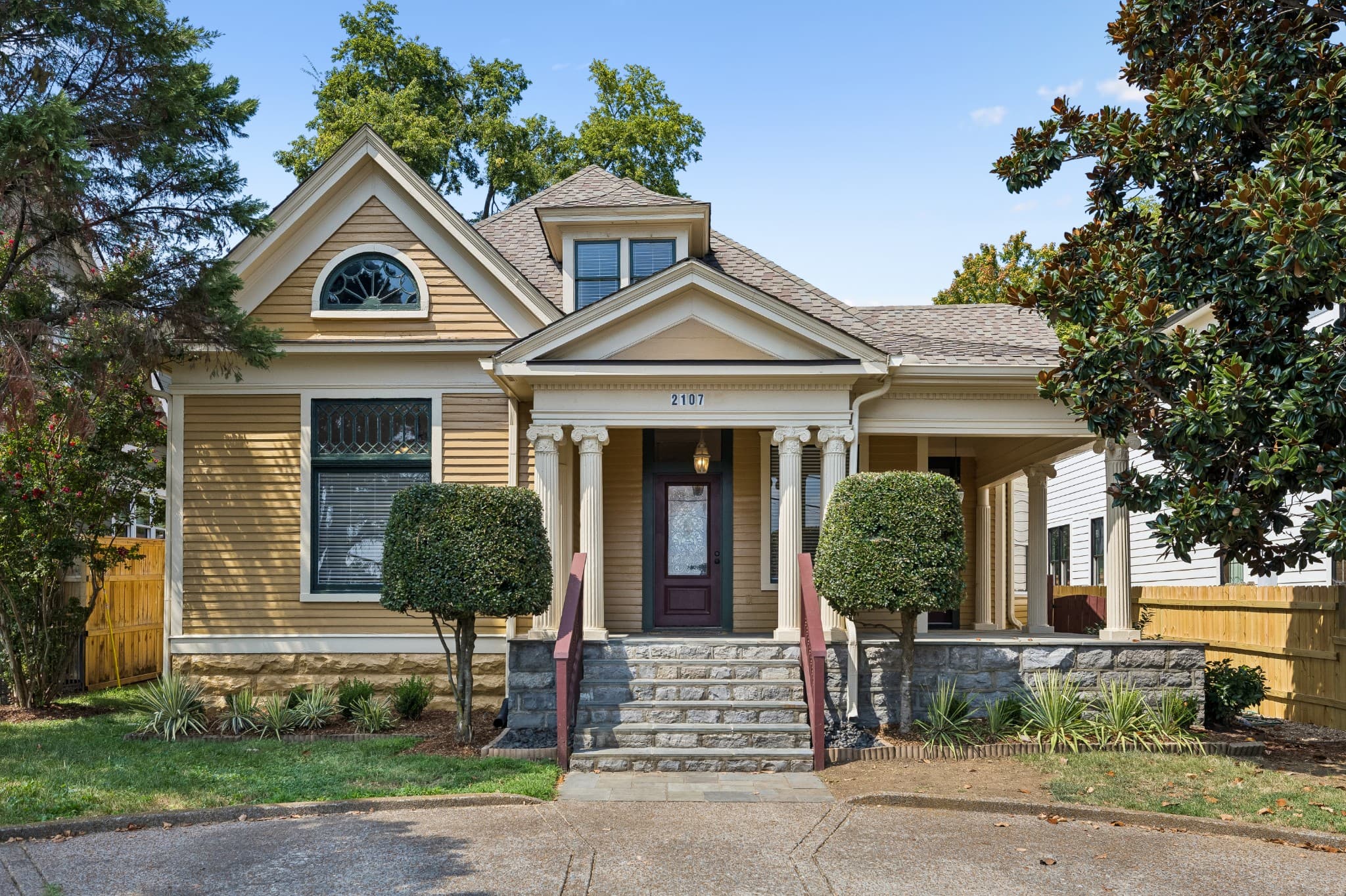 Front exterior of 2107 10th Avenue South, a yellow two story 1930 home with a front gable, an arched attic window, a covered front porch with columns and stone steps, one of the 12 South homes for sale in Nashville