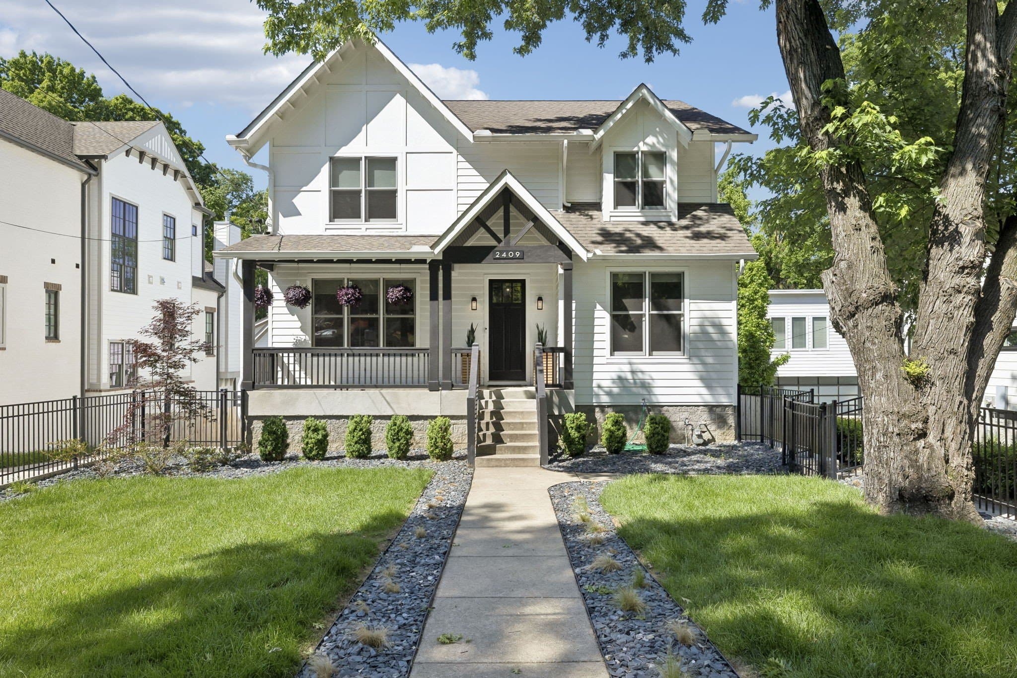 Front exterior of 2409 Vaulx Lane, a white two story home with a concrete walkway, black metal front fence, hanging porch baskets and the house number 2409 above the door, one of the 12 South homes for sale in Nashville