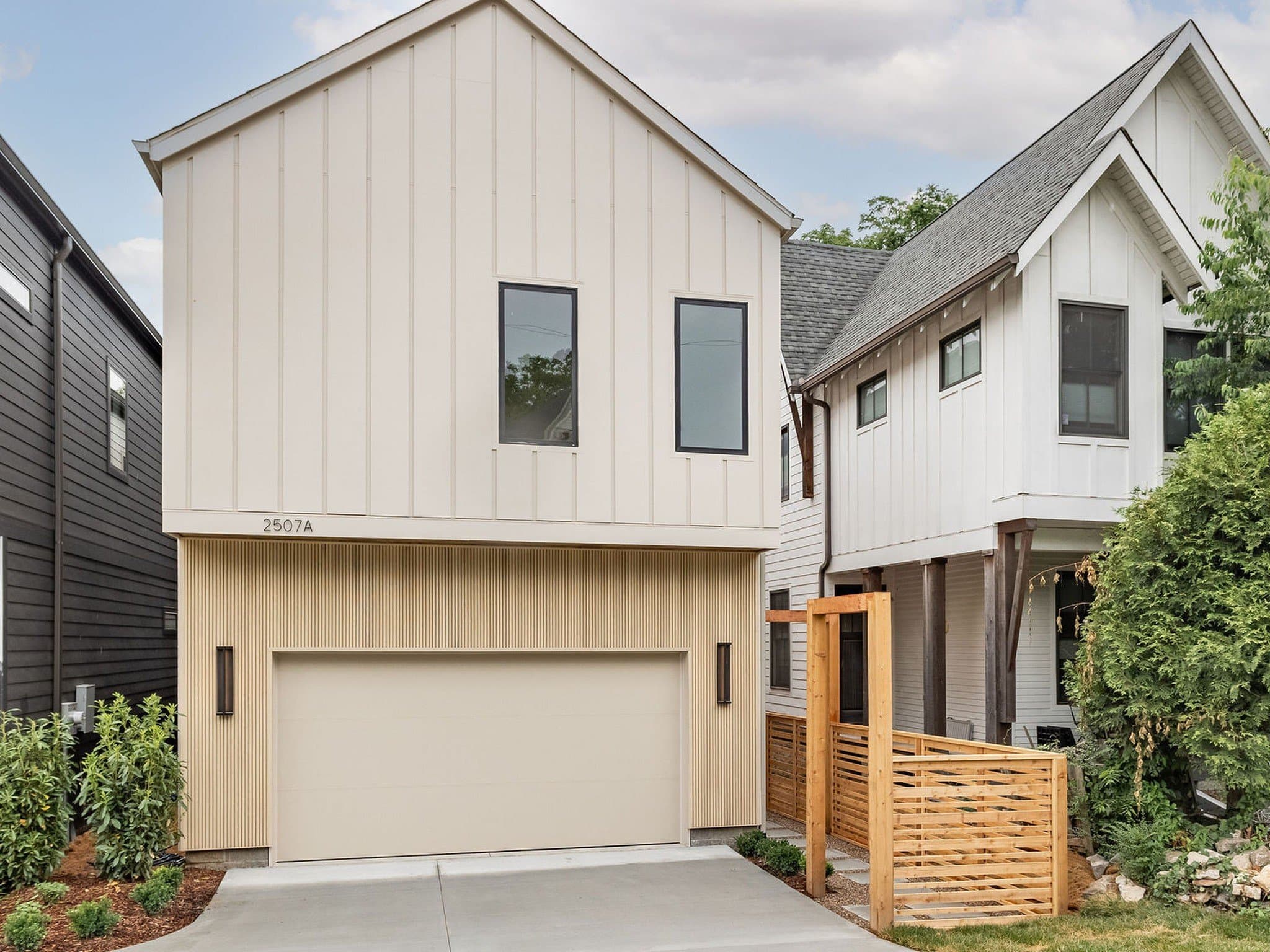 Street view of the front exterior of 2507A Vaulx Lane, a cream Contemporary two story home with vertical board and batten siding, a two car garage, the house number 2507A on the upper wall, and a wood fence and gate beside the shared driveway, one of the 12 South homes for sale in Nashville