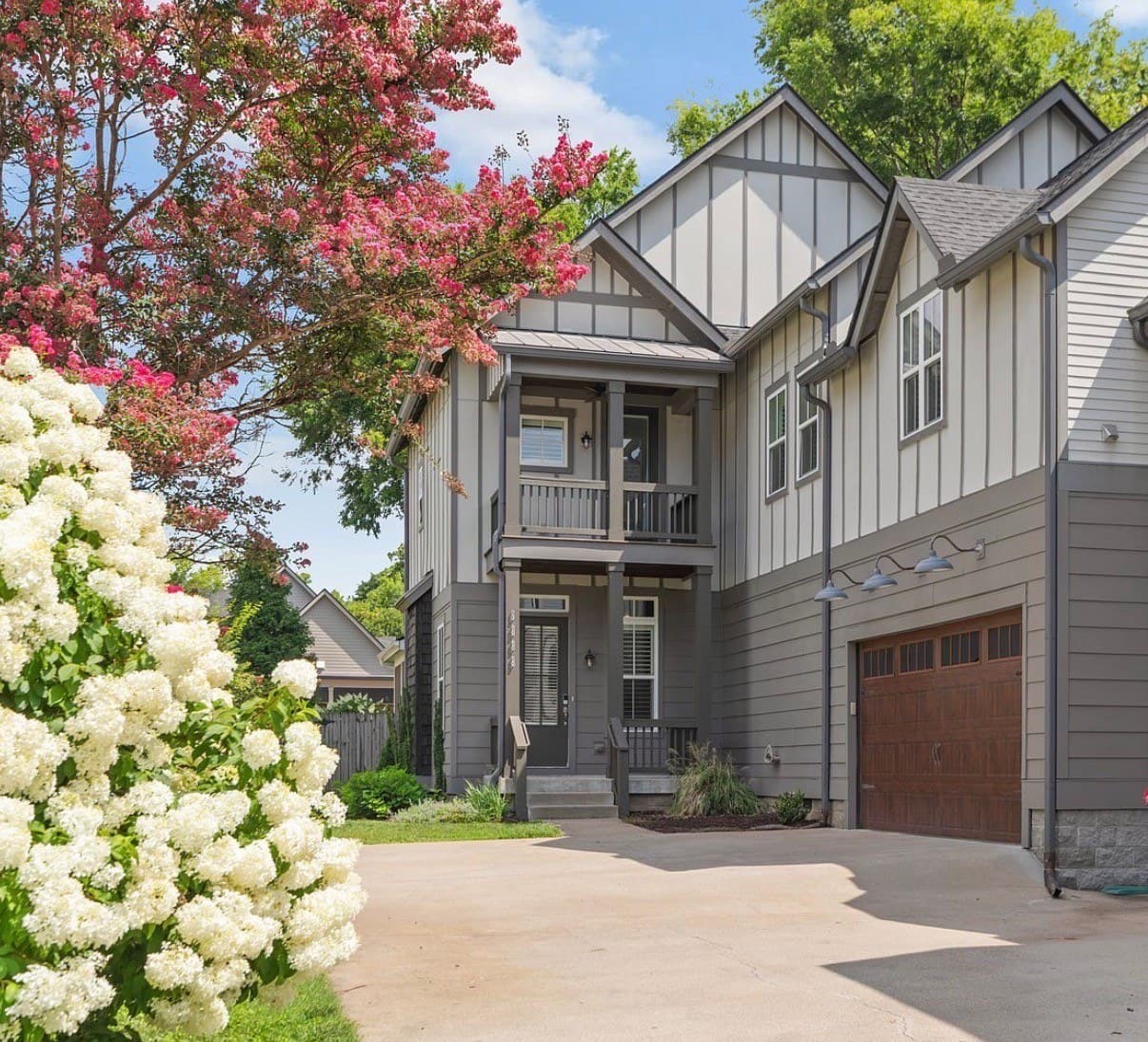 Driveway view of the front exterior of 908B Gale Lane, a gray board and batten two story home with a covered front porch, a balcony above it, a wood garage door and a concrete driveway framed by white hydrangeas, one of the 12 South homes for sale in Nashville